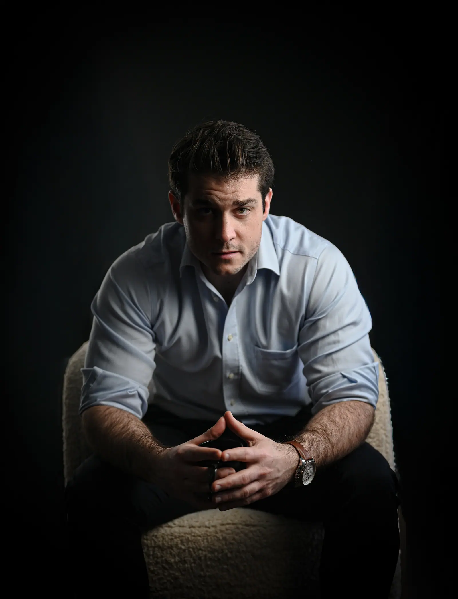 Man in a white shirt, seated, hands clasped, lit by a single source
