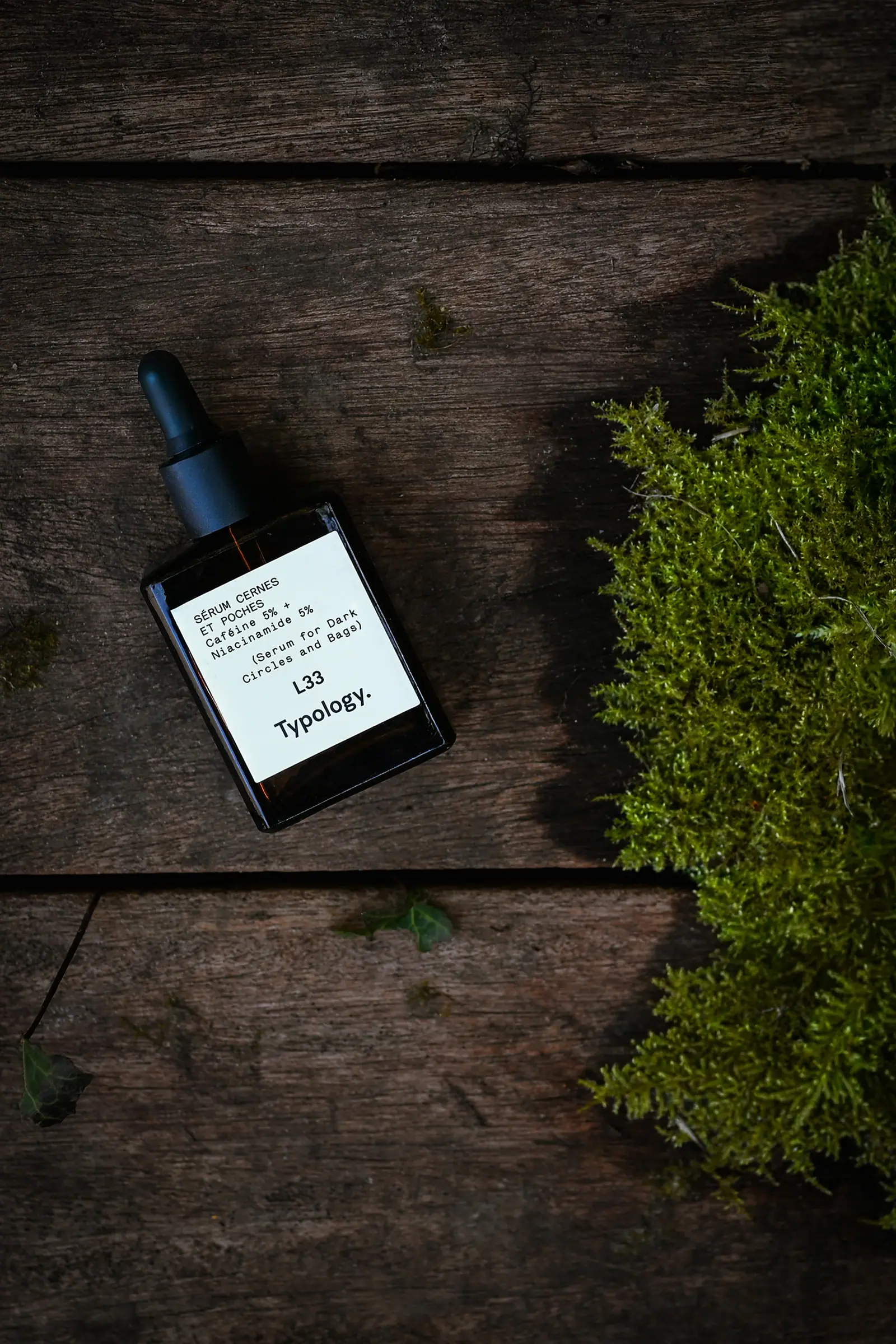 Typology serum on dark wood beside moss, seen from above